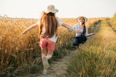 Kafasında şapkasıyla küçük mutlu bir kız annesine koşuyor buğday tarlasının yanında
