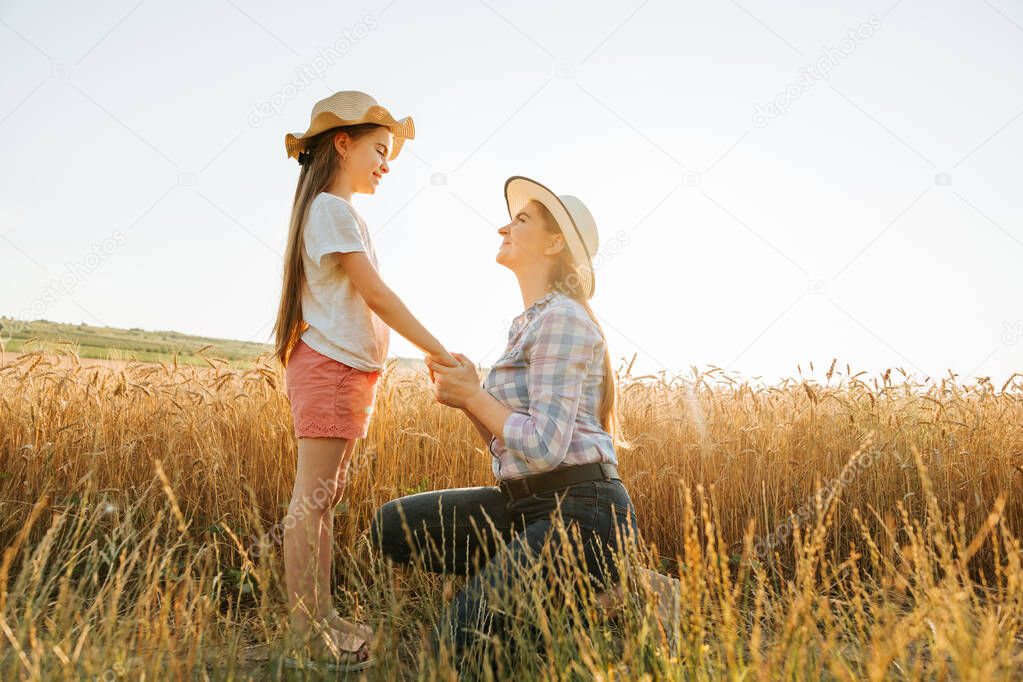 side view of mother and daughter in hat holding hands in golden wheat field.