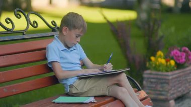 Mavi polo giyen genç okul çocuğu güneşli bir günde bacak bacak bacak üstüne atarak ders yazıyor. Sırt çantası yakında, arkada yeşil ağaçlar. Sakin, odaklanmış açık hava öğrenme anı.