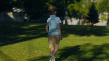 Polo tişörtlü ve şortlu neşeli genç bir çocuk sıcak bir günde enerji ve neşe dolu okulunun dışındaki güneşli çimenli bir alanda koşuyor..