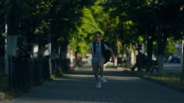 Yaz okulu kıyafetli genç bir çocuk aydınlık bir günde ağaçlı kaldırımdan aşağı koşar. Sırt çantası aceleyle giderken zıplıyor, enerji ve iyi hislerle dolu..