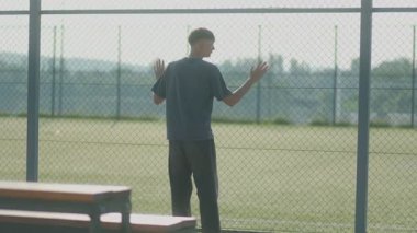 Genç bir çocuk çitlerin arkasında sessizce duruyor, boş bir spor sahasına bakıyor. Parlak gün ışığı, düşünceli ve sessiz duyguları arttırır..