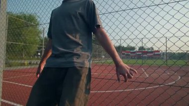 A teenager leans back against a sports fence, looking serious and focused, capturing the contemplative side of youth after practice on a sunny day.