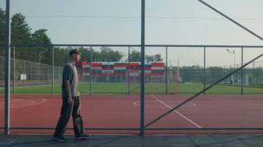 Genç adam sokak giysisi tarzında kırmızı bir kortun yanına eğiliyor. Çitlerde gölgeler, arka planda ağaçlar olan soğuk bir yaz havası. Sessiz Amerikan anı.