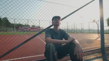 Teen boy sits near a fenced basketball court, bathed in sunset light, deep in emotional reflection. Scene expresses solitude, melancholy, and growing pains.