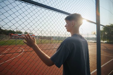 Gençler çitlere dokunuyor, gözler boş kortta, gün batımının yumuşak parıltısıyla parlıyor. Okul bahçesinde bir dakikalık sessizlik ve derinlik..