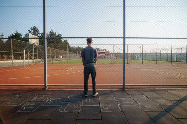 Çitlere bakan gençler, güneşin gölgeleri tebeşirle işaretlediği geçmiş çocukluğun sessiz sembolleri ve zamanda kaybolan anların altında hareketsiz duruyorlar..
