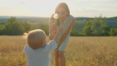 Şefkatli bir anne, bebeğine kırsalda güneşli bir günde karahindiba pufunu gösterir. Doğada huzurlu ve neşeli bir anı paylaşır..