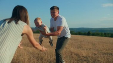 Neşeli bir aile, güneşli bir günde açık bir arazide yürümekte olan çocuklarıyla oynar. Bebek sıcak meltemde anne ve babasının arasında yükselirken neşeyle güler..