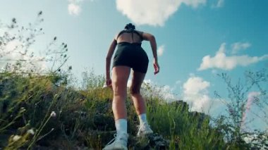 Aktif kıyafetli genç bir kadın güneşli gökyüzünün altında zorlu ve kayalık bir patikada koşuyor. Arka görünüşü, çabasını, enerjisini ve zirveye ulaşma isteğini gösteriyor..