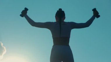 Zindelik düşkünü kadın temiz havada çalışıyor, halterleri kendine güvenle tutuyor. Pembe elbisesi arkasında bulutlar süzülürken patlıyor açık gökyüzünde.