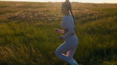 Pembe spor kıyafetli kadın altın saatinde açık arazide koşuyor. Uzun örgü ve doğal günbatımı harmanlama hareketi, sağlık ve sessiz güç.