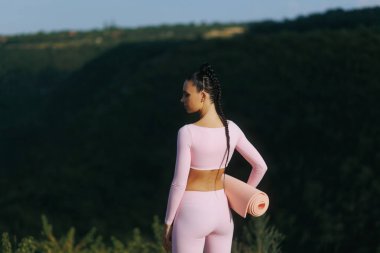 Pembe spor takım elbiseli, elinde yoga minderiyle zinde bir genç kadının arka görüntüsü. Açık havada, kendinden emin ve sakin, temiz dağ havasında eğitime ya da serinlemeye hazır..