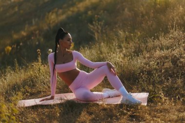 Pembe bir paspasın üzerinde oturan bu atletik kadın doğada sakin bir farkındalık uyguluyor. Vücudu hareketsiz ve güçlü. Dışarıyla bağlantı kurarken gözleri kapalı..