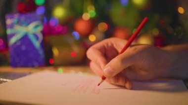 Kırmızı kalemli tanınmaz noel adamı bir mektup yazar. Bulanık arka plan, hediyeler, çelenk ışıkları. Seçili odak.