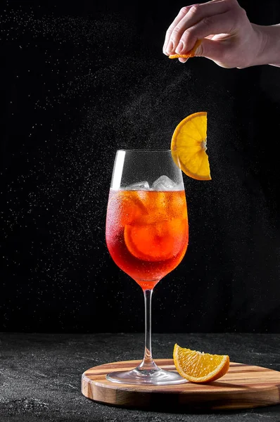 Aperol Spritz Cocktail em copo de vinho em tábua de madeira sobre fundo escuro. Mulher mão aperta casca de laranja — Fotografia de Stock