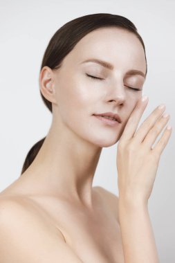 Güzel, sağlıklı bir yüzü olan genç bir kadın. Beyaz arka planda izole edilmiş. Stüdyoda üst düzey çekim, cilt bakımı konsepti. Eller ve parmaklar nazikçe yüz derisine dokunur, kahverengi saçlar ayrılır, kısa at kuyruğu içinde toplanır.