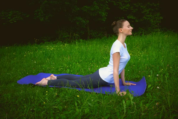 Профиль улыбающейся красивой молодой женщины, тренирующейся на синем коврике в аллее парка, делающей упражнения на растяжку — стоковое фото