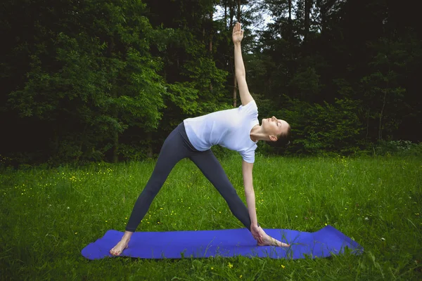 Yoga utthita trikonasana driehoek vormen door vrouw wit kostuum dragen op groen gras in het park rond pijnbomen — Stockfoto