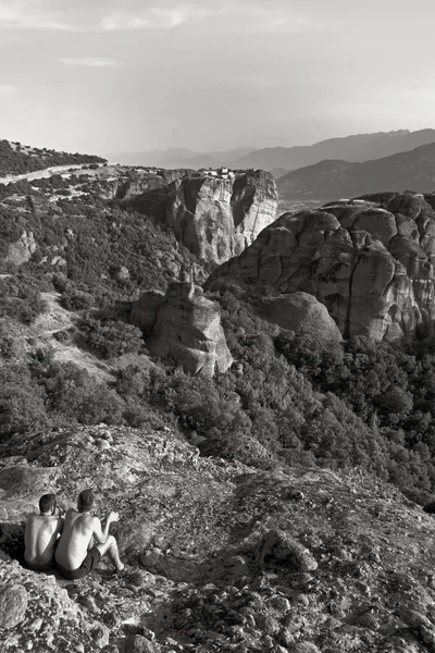 Meteora, Grécia. Paisagem de montanha com dois turistas sentados em rochas Meteora e Mosteiro de Roussanou, local de paisagem de mosteiros na rocha — Fotografia de Stock