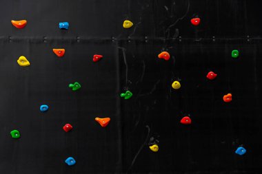 Çocuk parkındaki siyah bir duvardaki kaya tırmanışçıları için çok renkli kancaların ön görüntüsü. Spor ekipmanlarıyla renkli soyut arkaplan