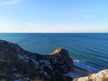 Vahşi bir taş plajında deniz dalgaları kayalara çarpar. Sakin deniz suyu. Mavi gökyüzünde beyaz bulutlar. Deniz dinlenmesi. Büyük taşlar. köpük.