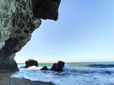 Vahşi bir taş plajında deniz dalgaları kayalara çarpar. Sakin deniz suyu. Mavi gökyüzünde beyaz bulutlar. Deniz dinlenmesi. Büyük taşlar. köpük.
