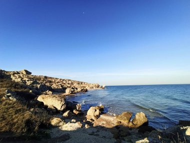 Vahşi bir taş plajında deniz dalgaları kayalara çarpar. Sakin deniz suyu. Mavi gökyüzünde beyaz bulutlar. Deniz dinlenmesi. Büyük taşlar. köpük.