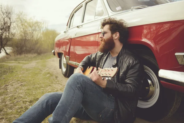 Barbudo hombre tocando la guitarra al aire libre cerca de coche retro — Foto de Stock