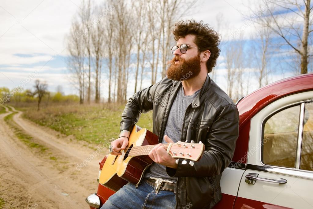Bearded man playing guitar outdoors near retro car