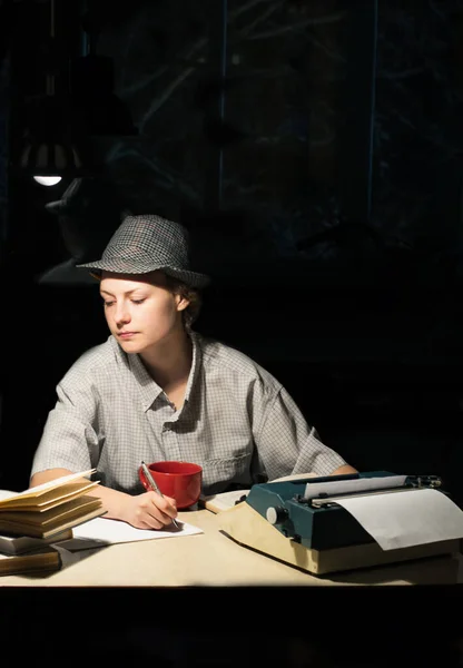 一个戴帽子的女孩坐在桌子旁 手里拿着打字机和书本 晚上做笔记的画像 — 图库照片