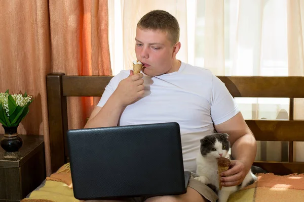 Man with laptop and cat eating ice cream cone in the bed