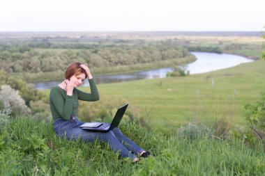 Güzel kız nehir kenarındaki çimlerin üzerinde grafik tabletini kullanıyor.