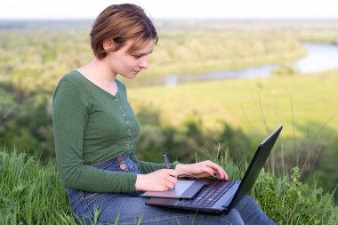 Güzel kız nehir kenarındaki çimlerin üzerinde grafik tabletini kullanıyor.