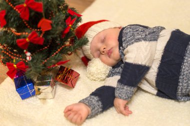 Noel Baba şapkalı küçük bebek Noel ağacının altında hediyelerle uyuyor.