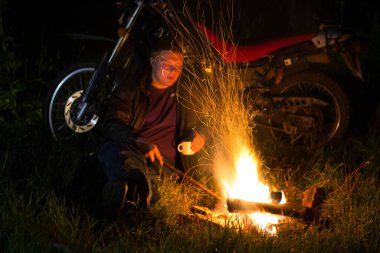 Adam şömine ve motosikletin yanında çay içip oturur.