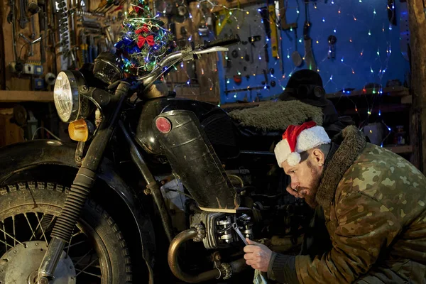 除夕之夜 一个头戴圣爪帽的男人在车库里修理一辆旧摩托车 — 图库照片