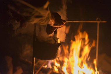 Hand of man taking the kettle, wich heated on a bright fire at night outdoor clipart