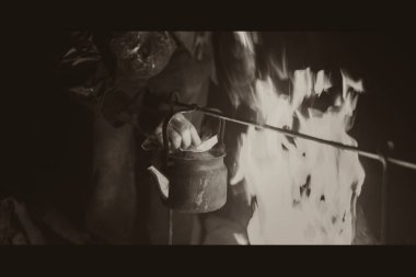 Gece açık havada parlak bir ateşte ısıtılan su ısıtıcısını alan adamın eli. Siyah beyaz fotoğraf.