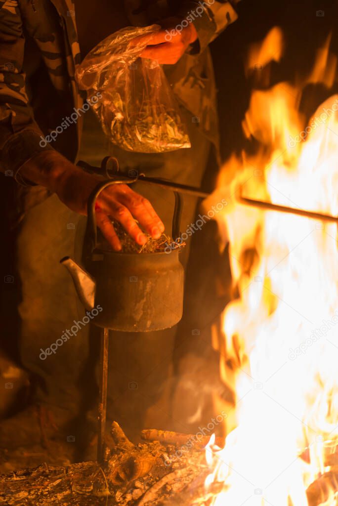 A man is laying dried herb in a kettle boiling on a bright fire at night