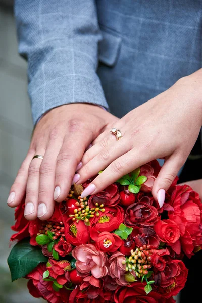 Newlyweds show hands with wedding rings on the fingers on a red bouquet, close-up.