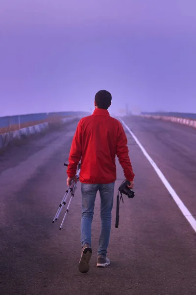 A man walks along an asphalt road with a camera and a tripod on an early foggy morning, back view.