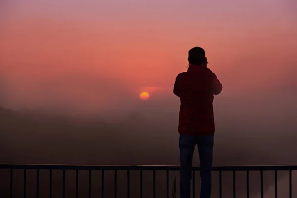 A silhouette of a man on a bridge over the river that photographs a sunrise on a foggy early morning.