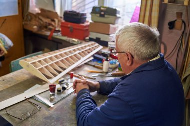 Kıdemli adam, atölyedeki masaüstünde uzaktan kumandalı bir uçağın ahşap kanadını yapıyor. Uçağın yapımı, arka görüş..