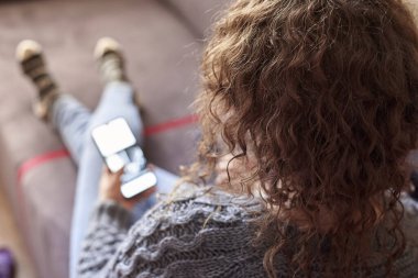 Kıvırcık saçlı bir kadın evdeki koltukta oturur ve cep telefonuna bakar..