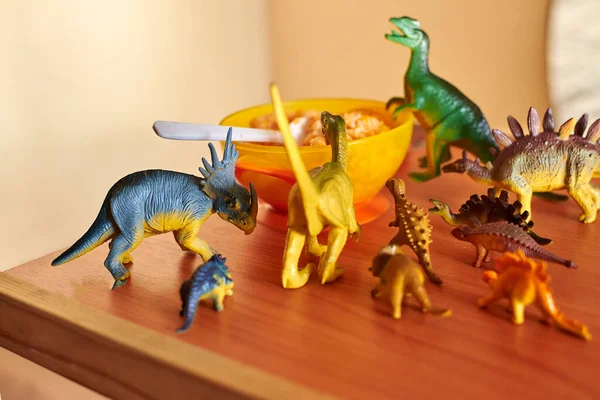 Toy dinosaurs stand on a children\'s table near a plate of porridge with spoon.