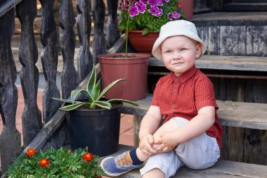 Küçük neşeli çocuk saksı çiçeklerinin yanındaki eski bir evin merdivenlerinde oturuyor..