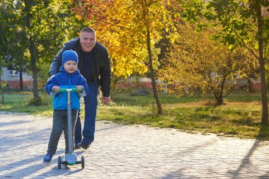 Babam iki yaşındaki oğluna sonbaharda parkta scooter sürmeyi öğretiyor..