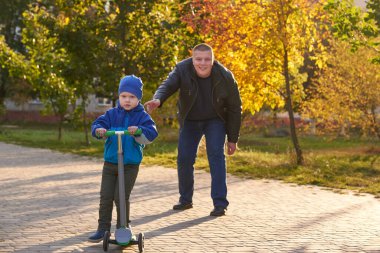 Babam iki yaşındaki oğluna sonbaharda parkta scooter sürmeyi öğretiyor..
