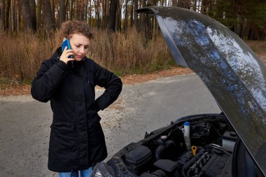 Üzgün bir kadın bozuk arabasının yanında duruyor ve sonbaharda yolda cep telefonuyla konuşuyor..